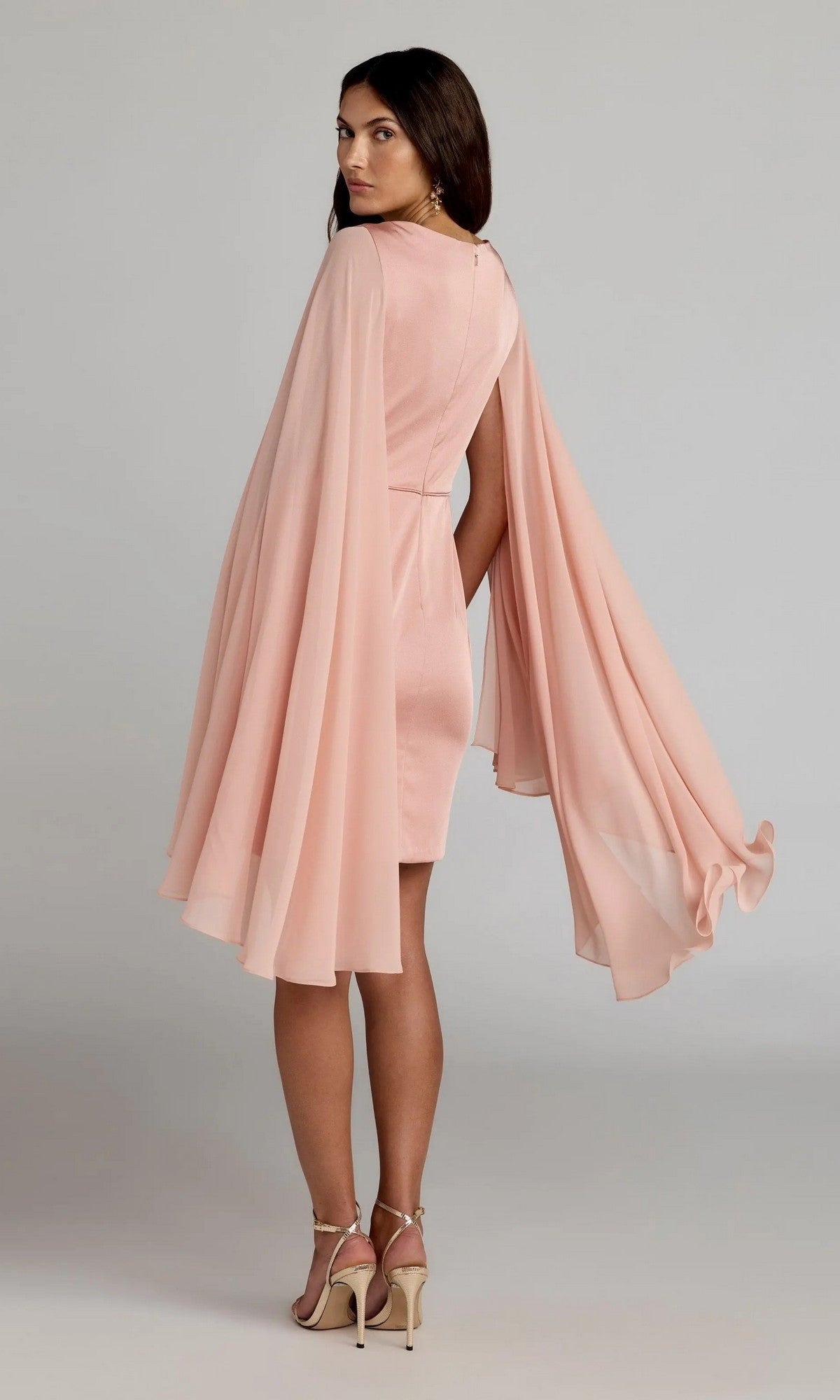 A woman models the Mac Duggal 55358 short semi-formal dress in light pink, featuring a V-neck and long chiffon cape sleeves, paired with high heels against a plain background.